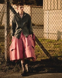 Young man leaning against a chain-link fence wearing a patterned shirt and pink cargo shorts