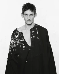 Portrait of a male model wearing a black embellished top adorned with small metallic pins against a white background