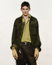 Model in olive green jacket, button-up shirt, and tan leather gloves against a plain background for The Greatest Magazine.