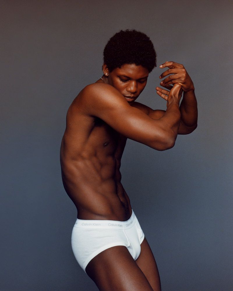 Portrait of a muscular shirtless Black man with an afro in white underwear, posing in front of a neutral grey backdrop indoors.