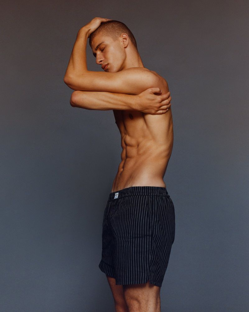 Profile shot of a young adult male with a shaved head, his torso exposed, wearing striped boxer shorts, posing against a grey background.