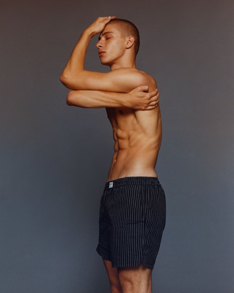 Striking portrait of a young, athletic male model in striped shorts with crossed arms and eyes closed against a neutral studio backdrop.
