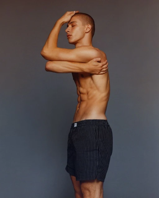 Striking portrait of a young, athletic male model in striped shorts with crossed arms and eyes closed against a neutral studio backdrop.