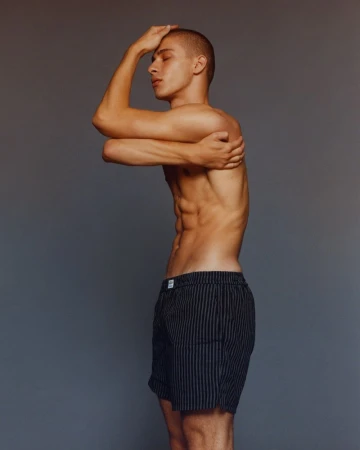 Striking portrait of a young, athletic male model in striped shorts with crossed arms and eyes closed against a neutral studio backdrop.