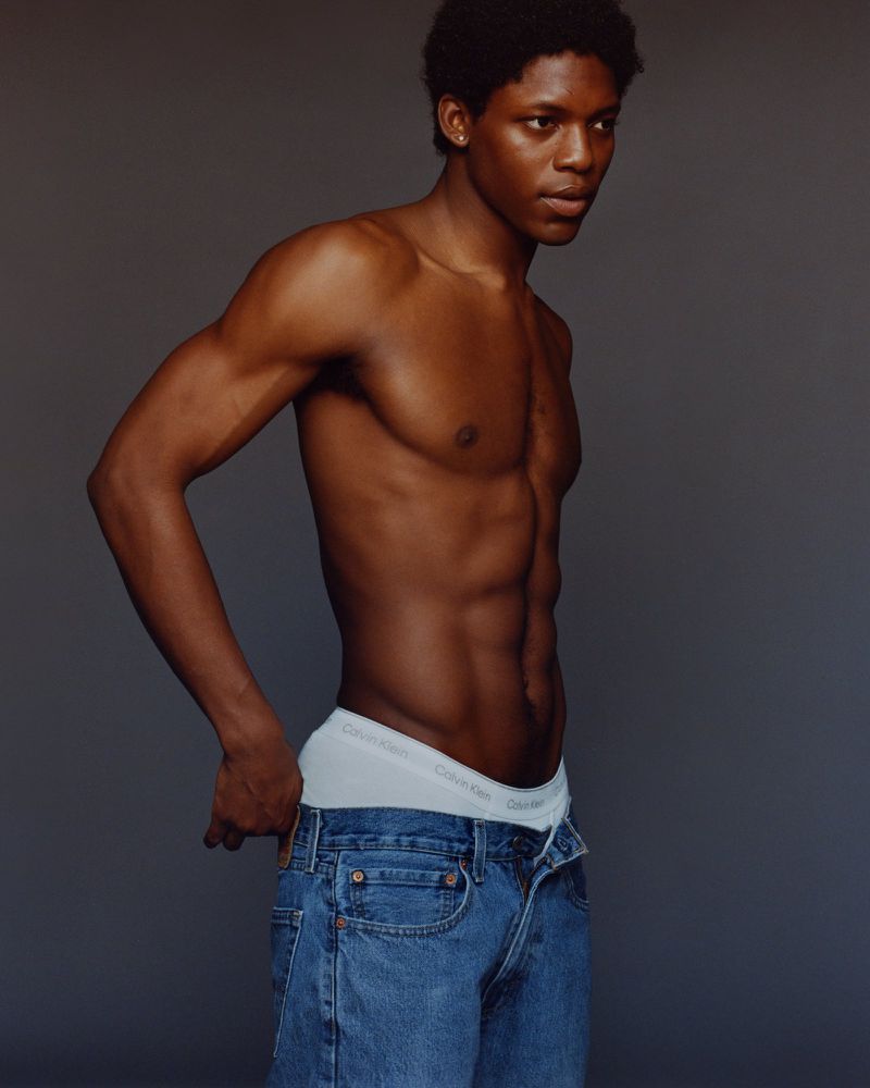 Portrait of a dark-skinned, shirtless man in jeans and underwear against a neutral backdrop, showing his physique.
