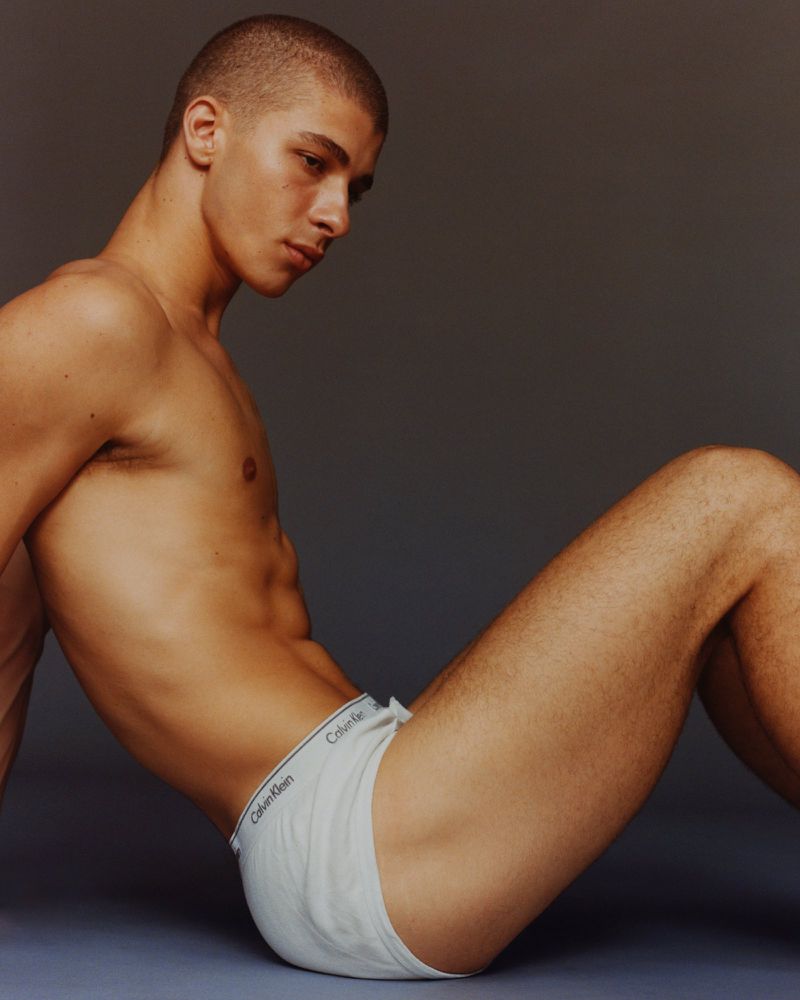 Man poses indoors wearing white Calvin Klein underwear. He has a buzzcut hairstyle and looks down while sitting against a grey wall.