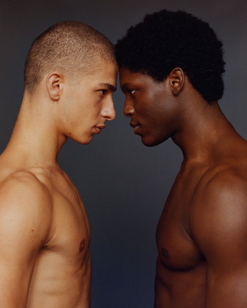 Portrait of two topless adult male models, one Black and one White, stand face-to-face against a gray background in a photo studio.
