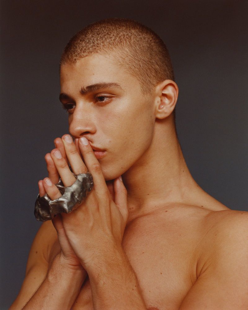 Portrait of a shirtless adult male with short hair wearing a silver ring, hands clasped together, gazing slightly to the left against a grey background.
