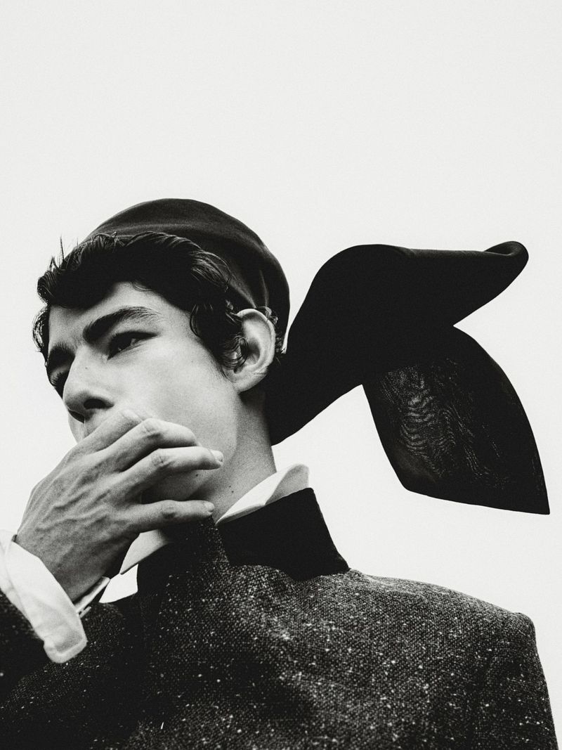 Black and white portrait of a person in vintage attire featuring a beret and textured jacket, face partially obscured by a hand.