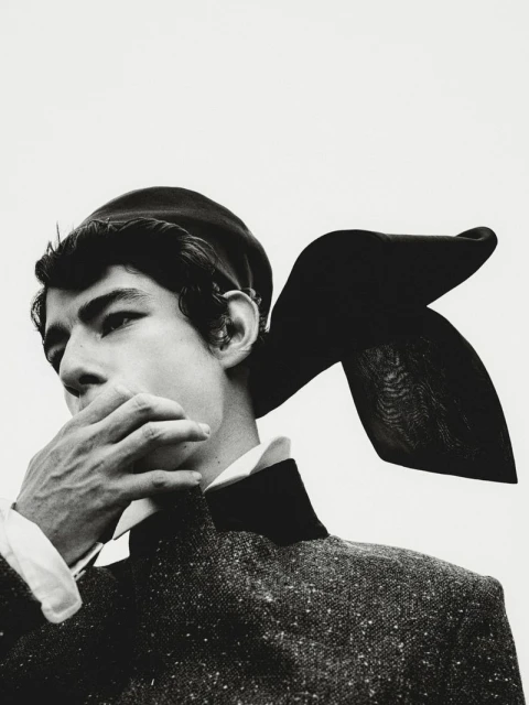Black and white portrait of a person in vintage attire featuring a beret and textured jacket, face partially obscured by a hand.