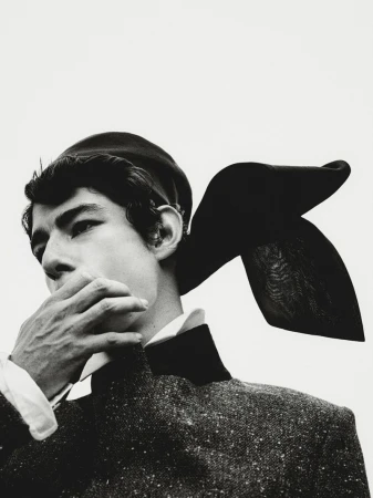 Black and white portrait of a person in vintage attire featuring a beret and textured jacket, face partially obscured by a hand.