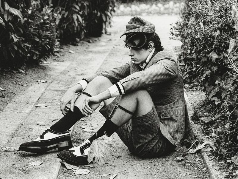 Monochrome portrait of a seated male model in tweed suit, cap, and spectator shoes, styled with garter clips outdoors on a stone path.