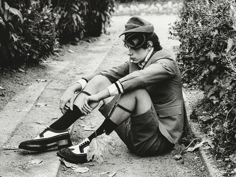 Monochrome portrait of a seated male model in tweed suit, cap, and spectator shoes, styled with garter clips outdoors on a stone path.