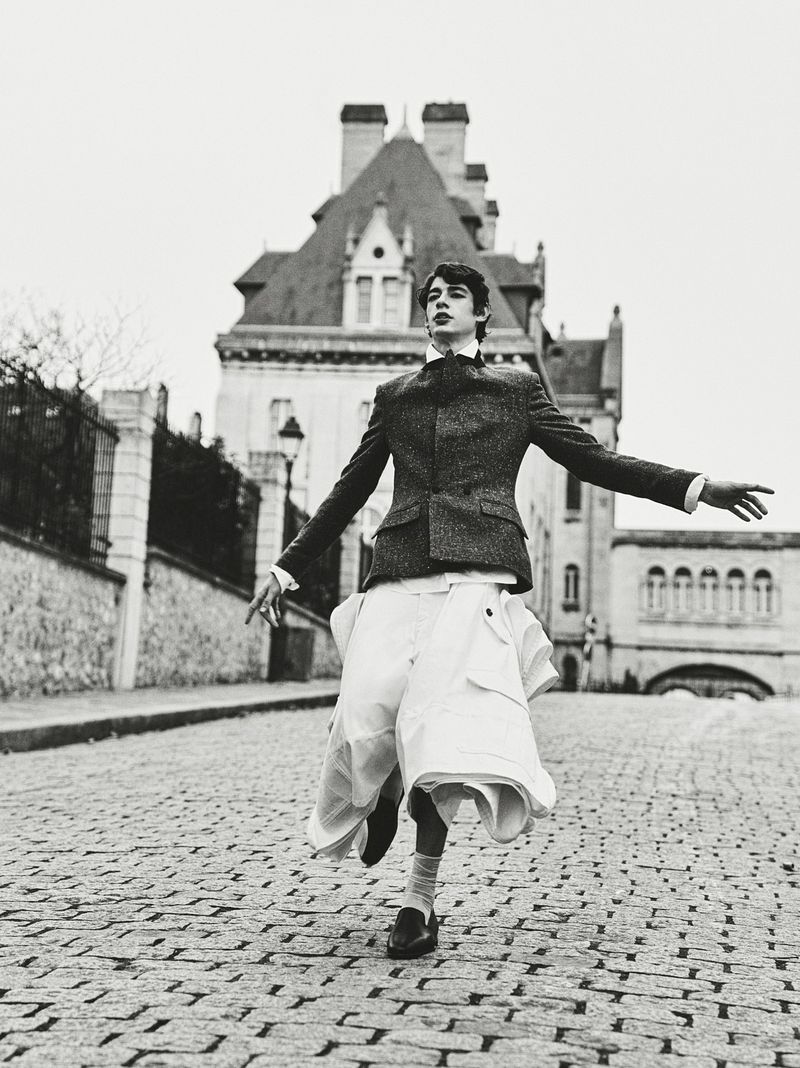 Young man in blazer and skirt strikes a playful pose on cobblestone street with building backdrop in a black and white editorial fashion shot.