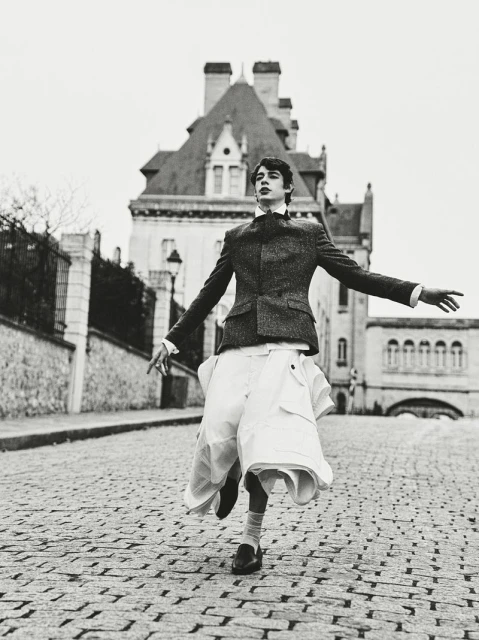 Young man in blazer and skirt strikes a playful pose on cobblestone street with building backdrop in a black and white editorial fashion shot.