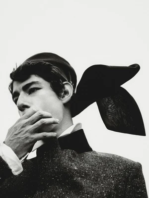 Close-up monochrome editorial portrait of a male model wearing a textured dark jacket with a cap and streaming ribbon against a pale background