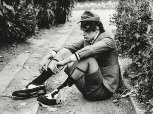 Model seated on a gravel path wearing a patterned blazer, shorts, stockings, and spectator shoes with a cap.