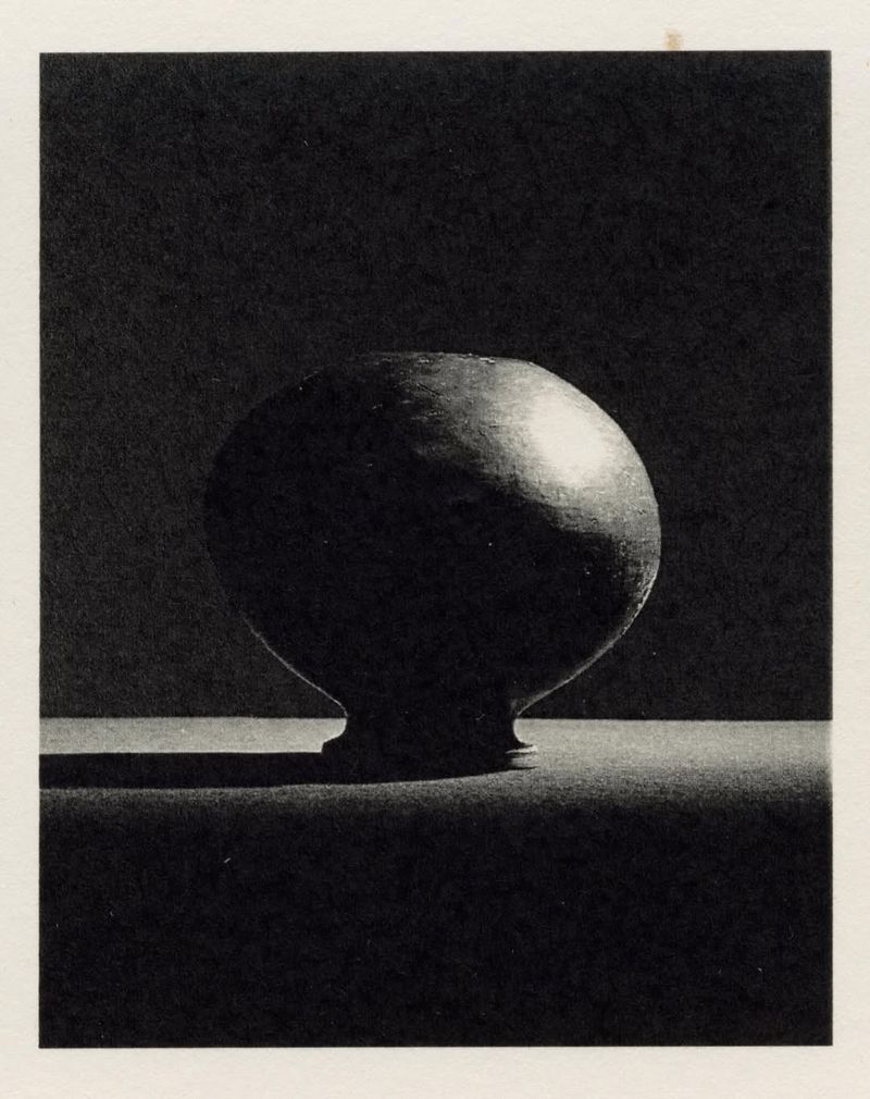 Monochromatic still life studio shot of round vase with foot, casting shadow against dark backdrop, showcasing texture and form.
