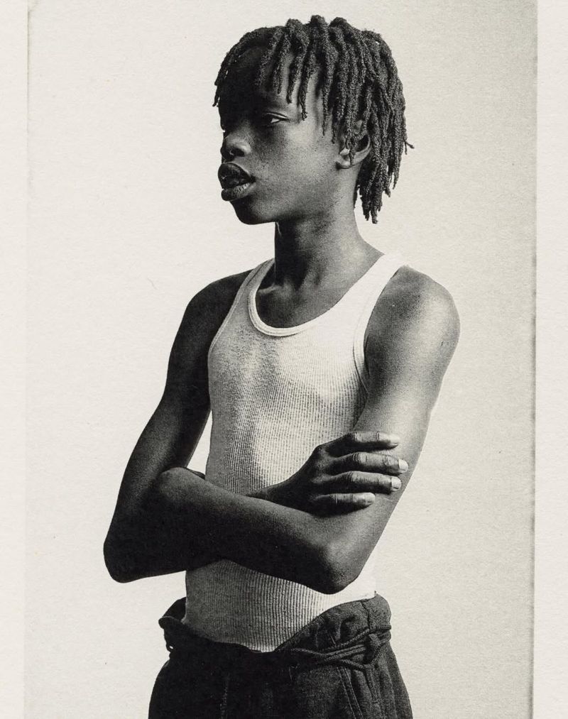 Monochrome portrait of a young person with dreadlocks wearing a tank top, arms crossed, looking off to the side in a neutral indoor setting.