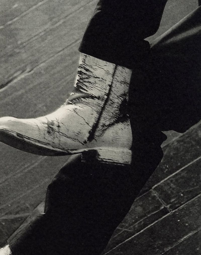 Close-up of a white cowboy boot with black distressed details, paired with dark trousers, set against a weathered wooden floor.