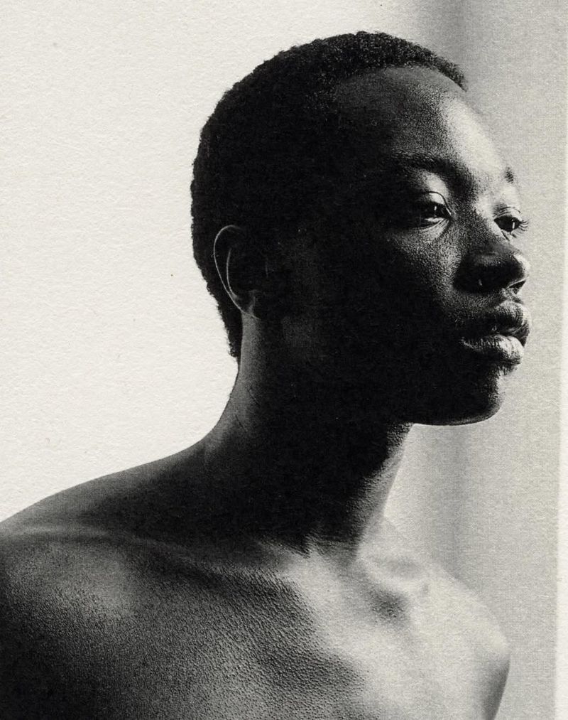 Monochrome portrait of a young Black man gazing to the right, showcasing his shoulders and upper chest against a light neutral background.