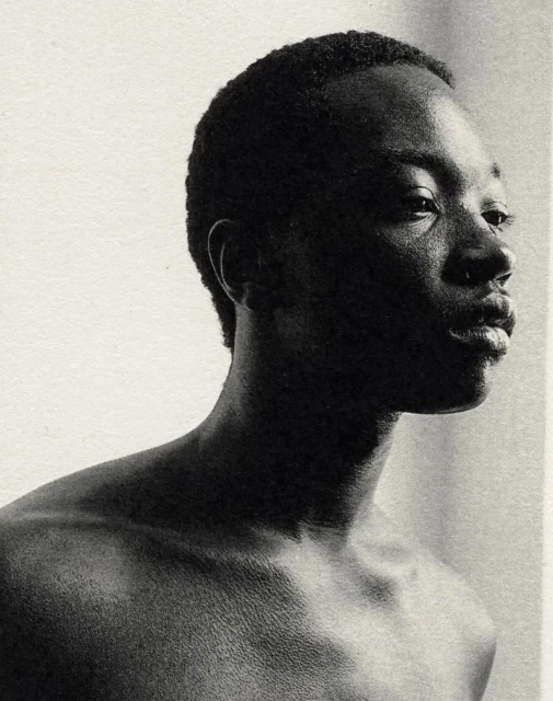 Monochrome portrait of a young Black man gazing to the right, showcasing his shoulders and upper chest against a light neutral background.