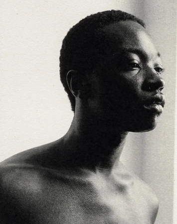Monochrome portrait of a young Black man gazing to the right, showcasing his shoulders and upper chest against a light neutral background.