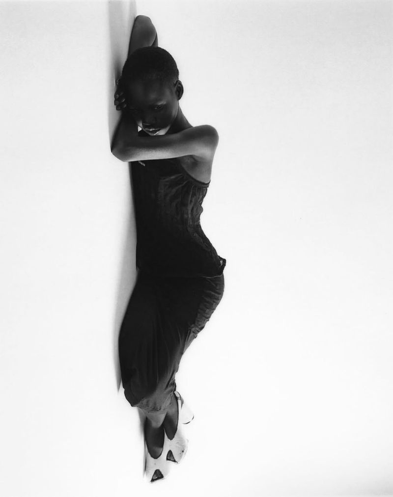 Black and white fashion shot of a slender adult female model leaning against a white wall, wearing a dress and pointed shoes.