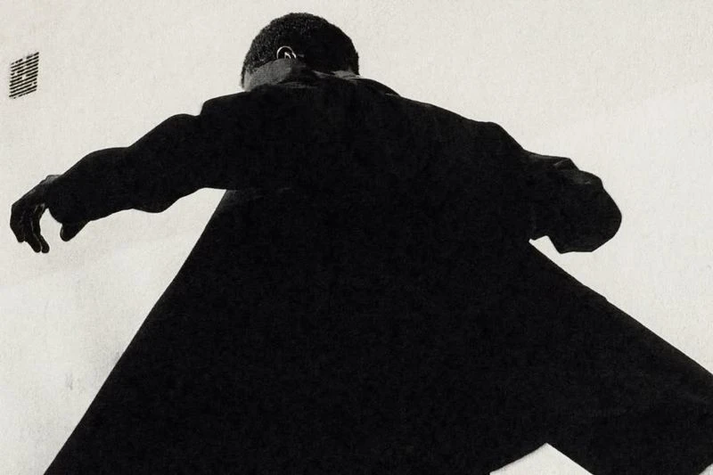 Low angle of a Black adult wearing a black coat, black gloves, and a partial view of his head against an off-white background.