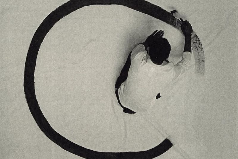 Overhead view of an artist in a white shirt using a long brush to paint a large black circle on a white canvas laid on the ground.