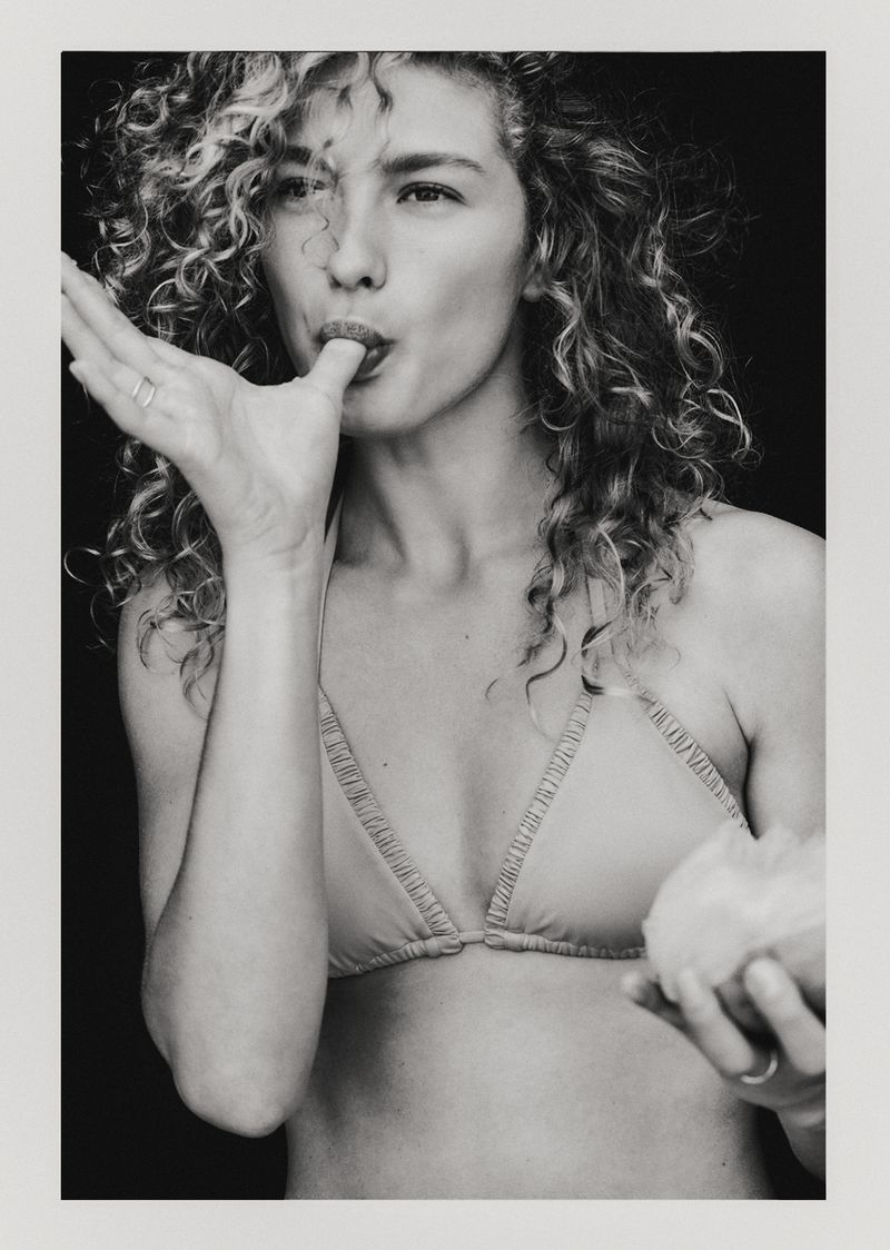 Monochrome portrait of a curly-haired woman wearing a bra top, licking her finger while holding a spherical fruit in her hand.