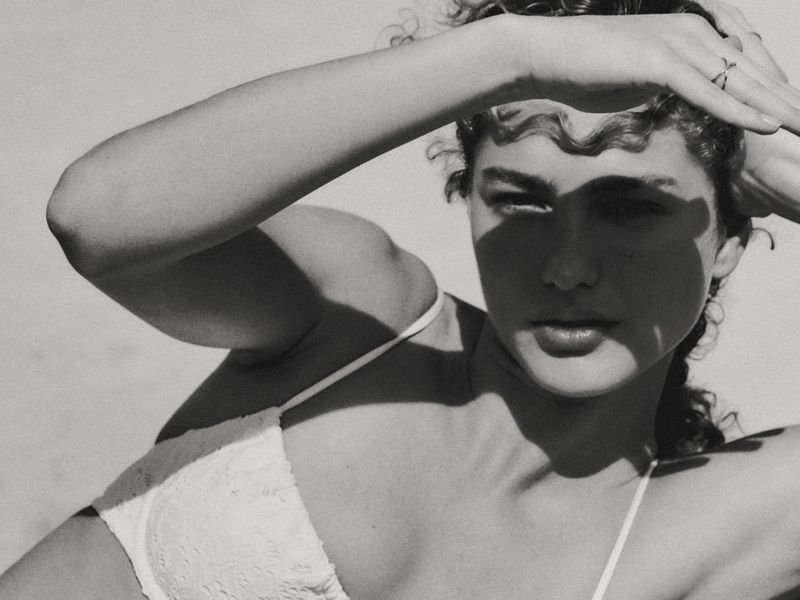 Black and white portrait of a young woman with curly hair shielding her face from the sun, wearing a lace bra top.