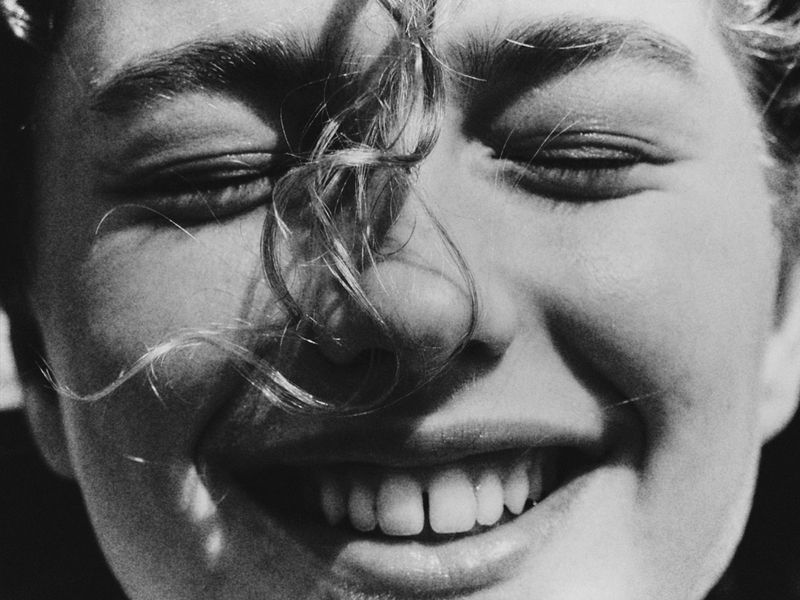 Monochrome close-up of a young adult female's face with her eyes closed, fair skin, light curly hair, and a natural smile in bright sunlight.
