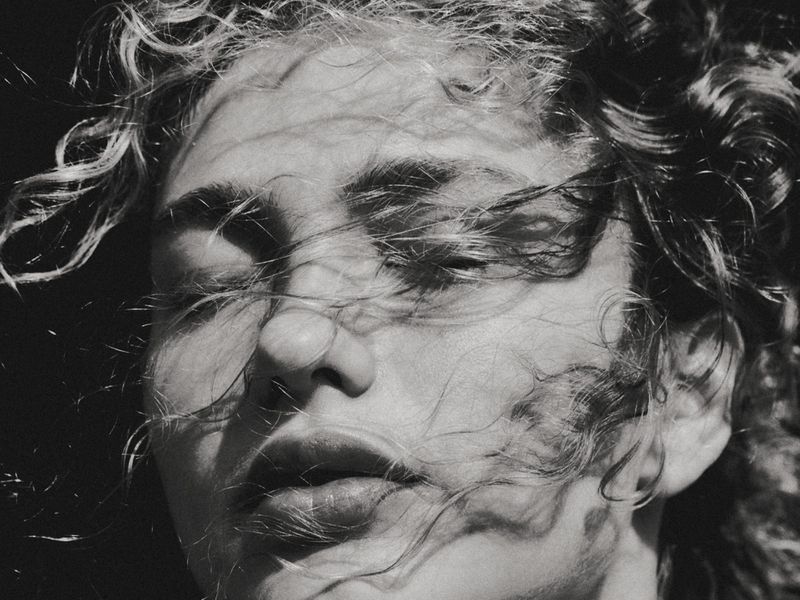 Black and white portrait of a young woman with curly hair, eyes closed, and hair blowing across her face in a natural outdoor setting.