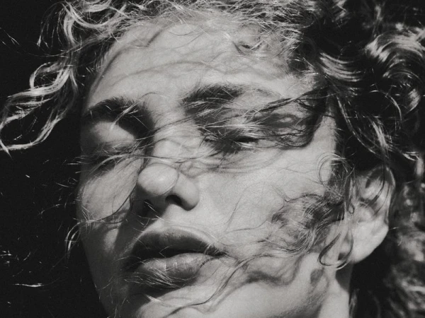 Black and white portrait of a young woman with curly hair, eyes closed, and hair blowing across her face in a natural outdoor setting.