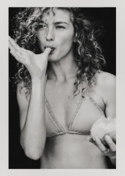 Black and white portrait of model Dorit Revelis wearing a bikini with curly hair and holding fruit against a dark background