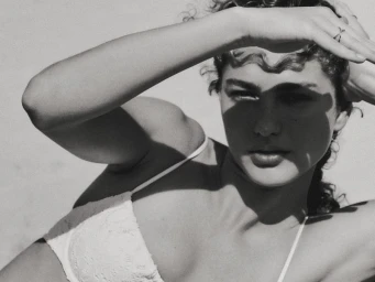 Model Dorit Revelis wearing a white bikini top, posing with her hand shielding her face against harsh sunlight in a black and white portrait.