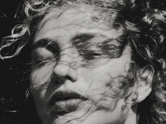 Close-up portrait of model Dorit Revelis with curly hair blowing across her face in black and white.