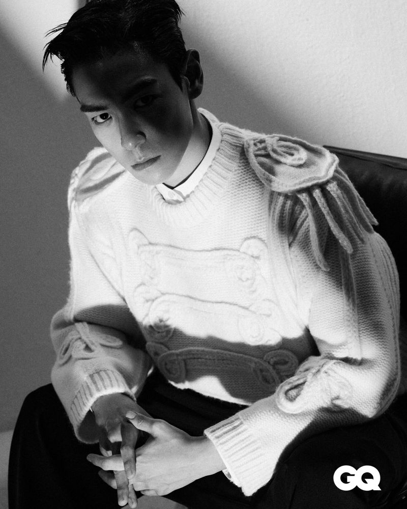 Black and white portrait of an Asian man in a patterned sweater sitting in a chair with his hands folded, cast in hard studio light.