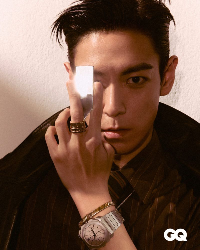 Close-up shot of an Asian man with slicked-back hair wearing a striped shirt, jacket, jewelry, and wristwatch indoors with soft lighting.