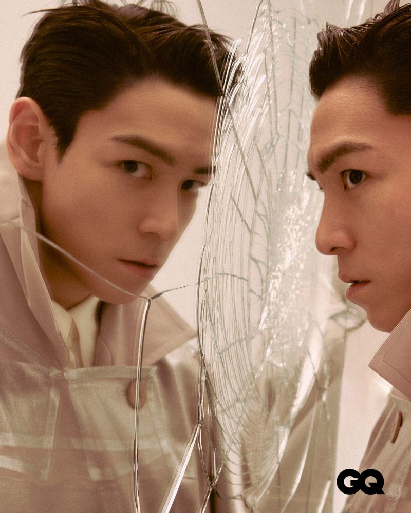 Portrait of an adult male looking in a shattered mirror wearing a light trench coat and collared shirt with a GQ logo visible.