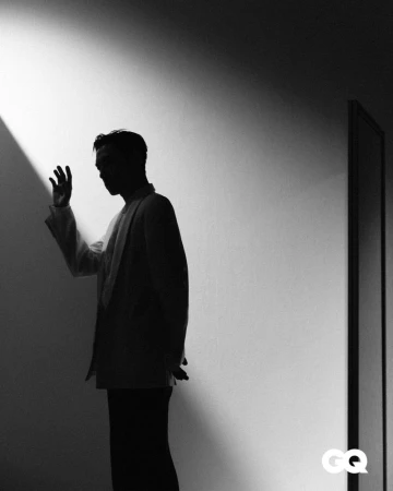 Monochromatic silhouette of a man in a suit jacket standing indoors against a wall with a beam of light, creating a high-contrast portrait.