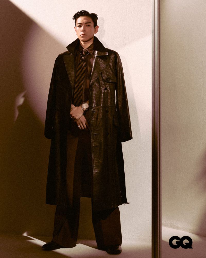 Stylish man in a dark trench coat poses indoors for a GQ magazine fashion editorial, complemented by a tie, wide-leg pants, and leather shoes.