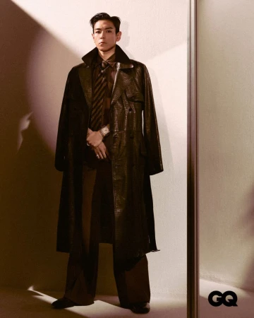 Stylish man in a dark trench coat poses indoors for a GQ magazine fashion editorial, complemented by a tie, wide-leg pants, and leather shoes.