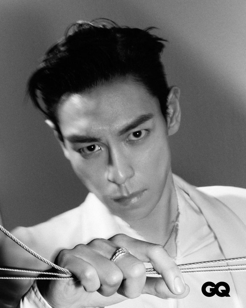 Black and white portrait of an adult male with dark hair, wearing a white shirt and rings, gazing downward and holding silver cords.
