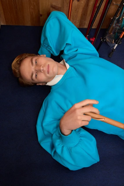 Young adult male model lying on navy carpet, wearing a teal blue sweater, white collar, holding a wooden cane, with skis in background.