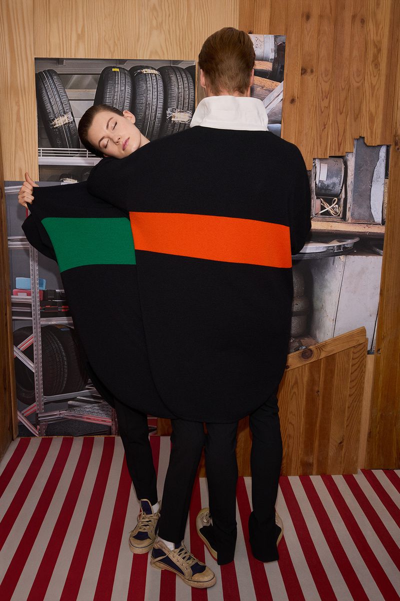 Two people in black sweaters stand in front of a wood paneled wall with a tire rack photo and striped carpet, fashion forward posing.