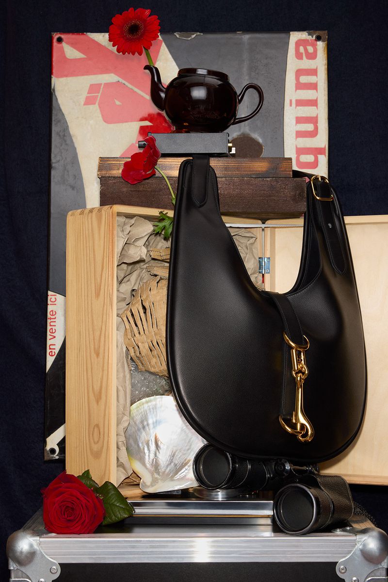 Black hobo bag with gold hardware displayed on wooden crates with flowers, shell, and vintage poster background.
