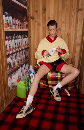 A woman sits on a plaid bag surrounded by cleaning supplies in a wood-paneled room with faux supermarket shelves in the background.