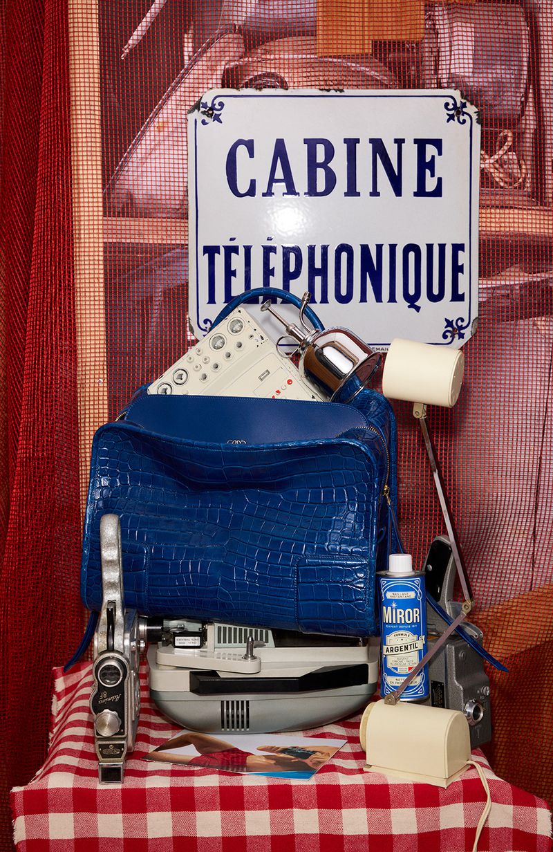 Retro display featuring a blue handbag, vintage cameras, cleaning supplies, and a French telephone booth sign on a checkered tablecloth.